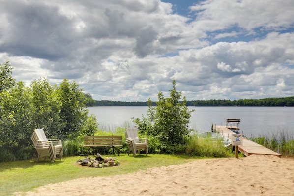 A lakeside setting with a wooden deck, lounge chairs, and a sandy area.