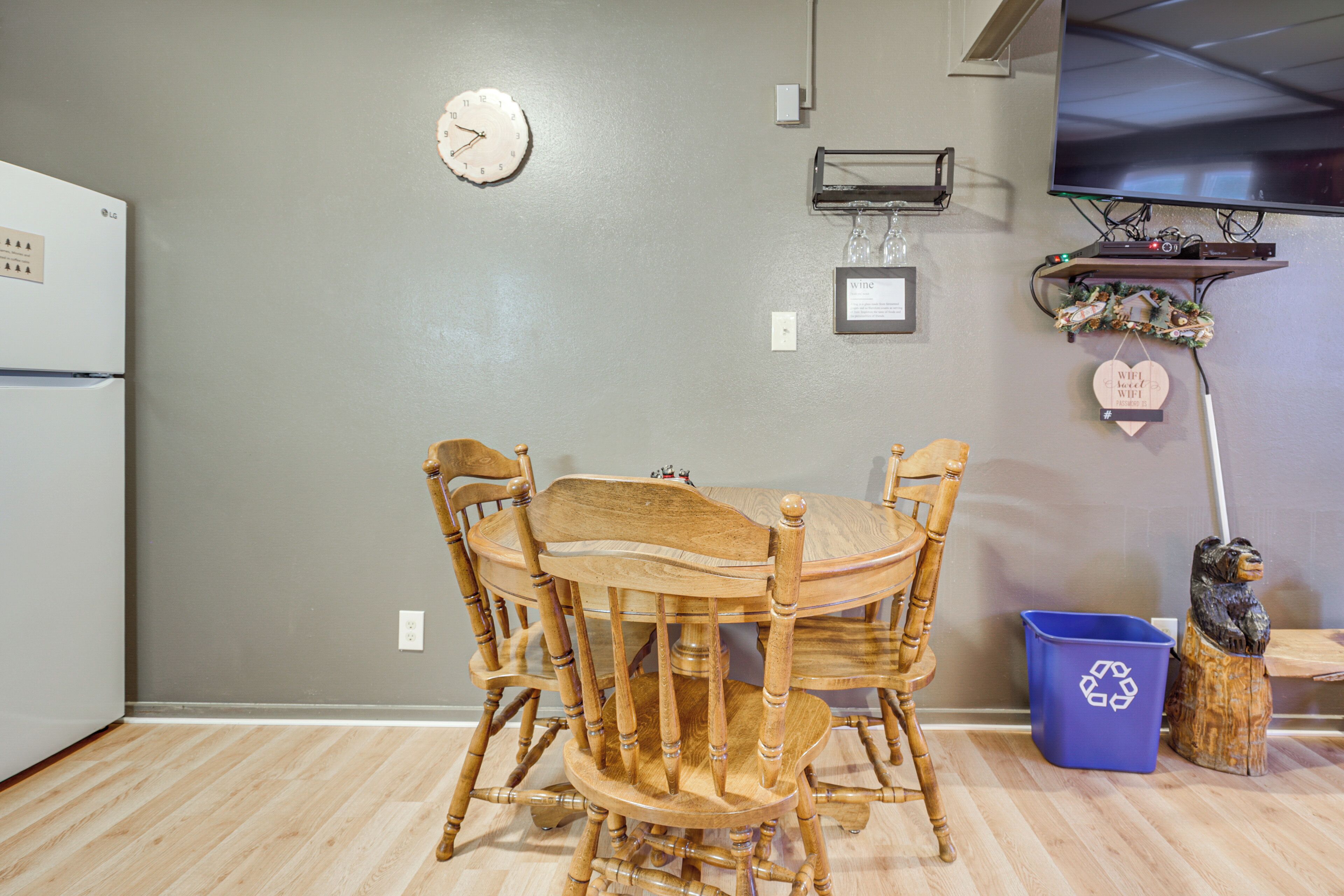 A kitchen with a dining table and chairs, a clock on the wall, a TV mounted above the counter, and a blue recycling bin.