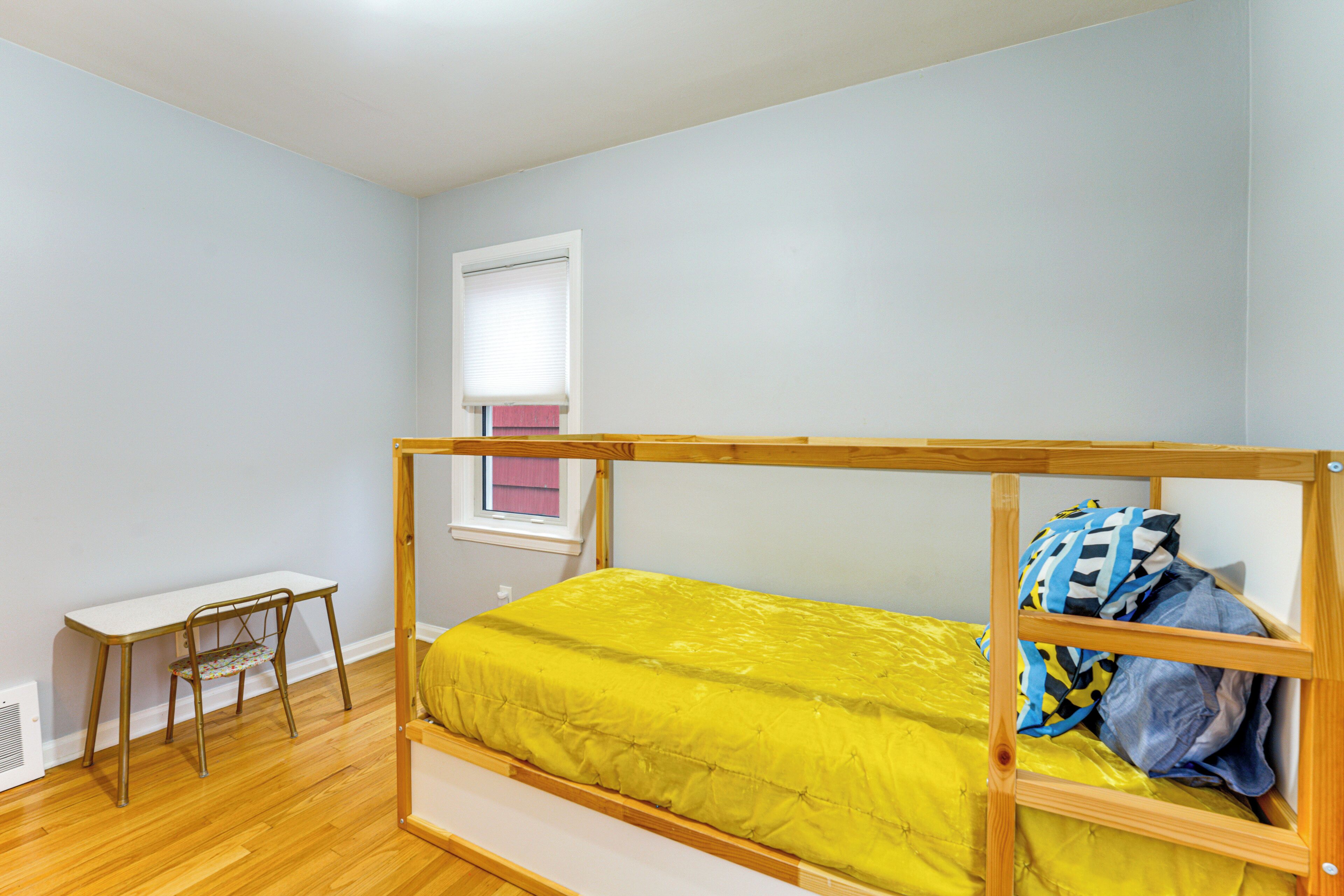 A single bed with a wooden frame and a yellow mattress, a small table with a chair, and a window with blinds.