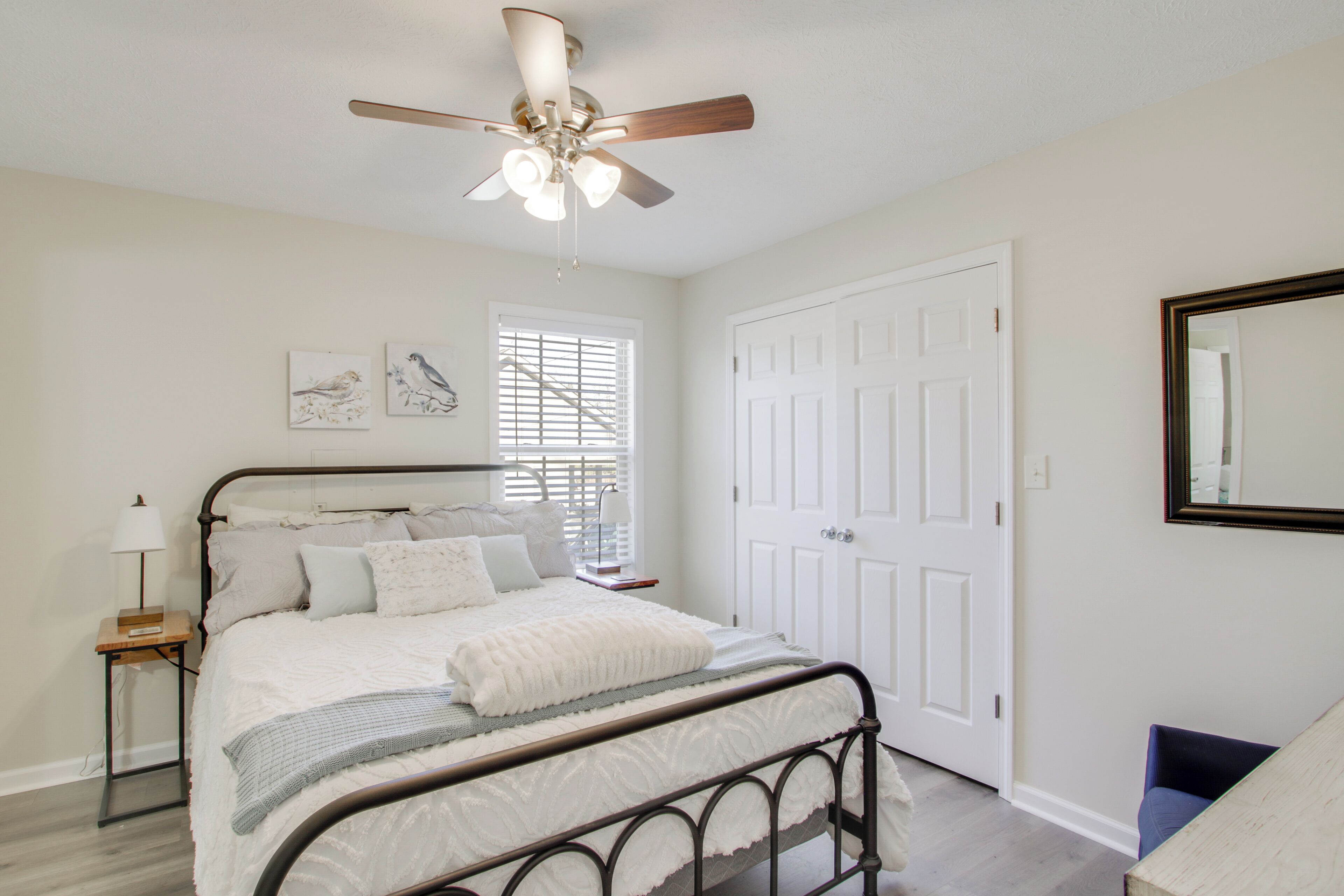 A bedroom with a bed, a ceiling fan, a window with blinds, a door, and a mirror on the wall.