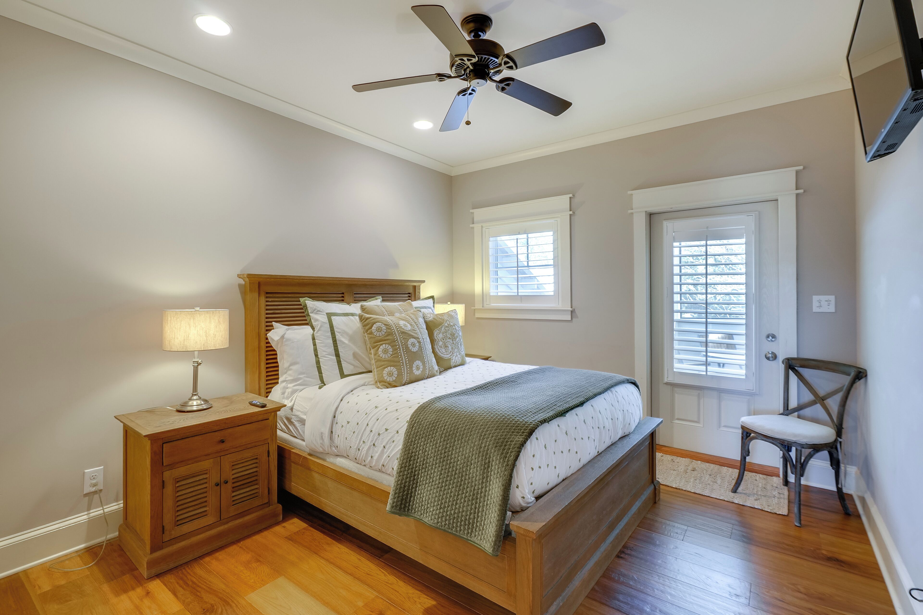 Une chambre à coucher avec un lit en bois, une table de chevet avec une lampe, un ventilateur de plafond, une chaise et une fenêtre avec des stores.
