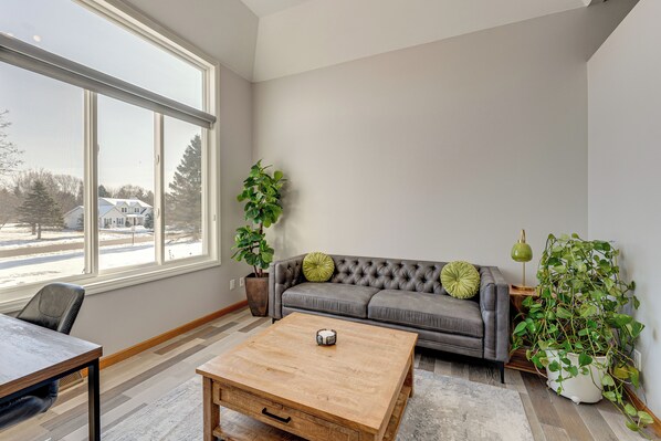 A modern living room with a grey sofa, a wooden coffee table, and a large window offering a view of a snowy street.