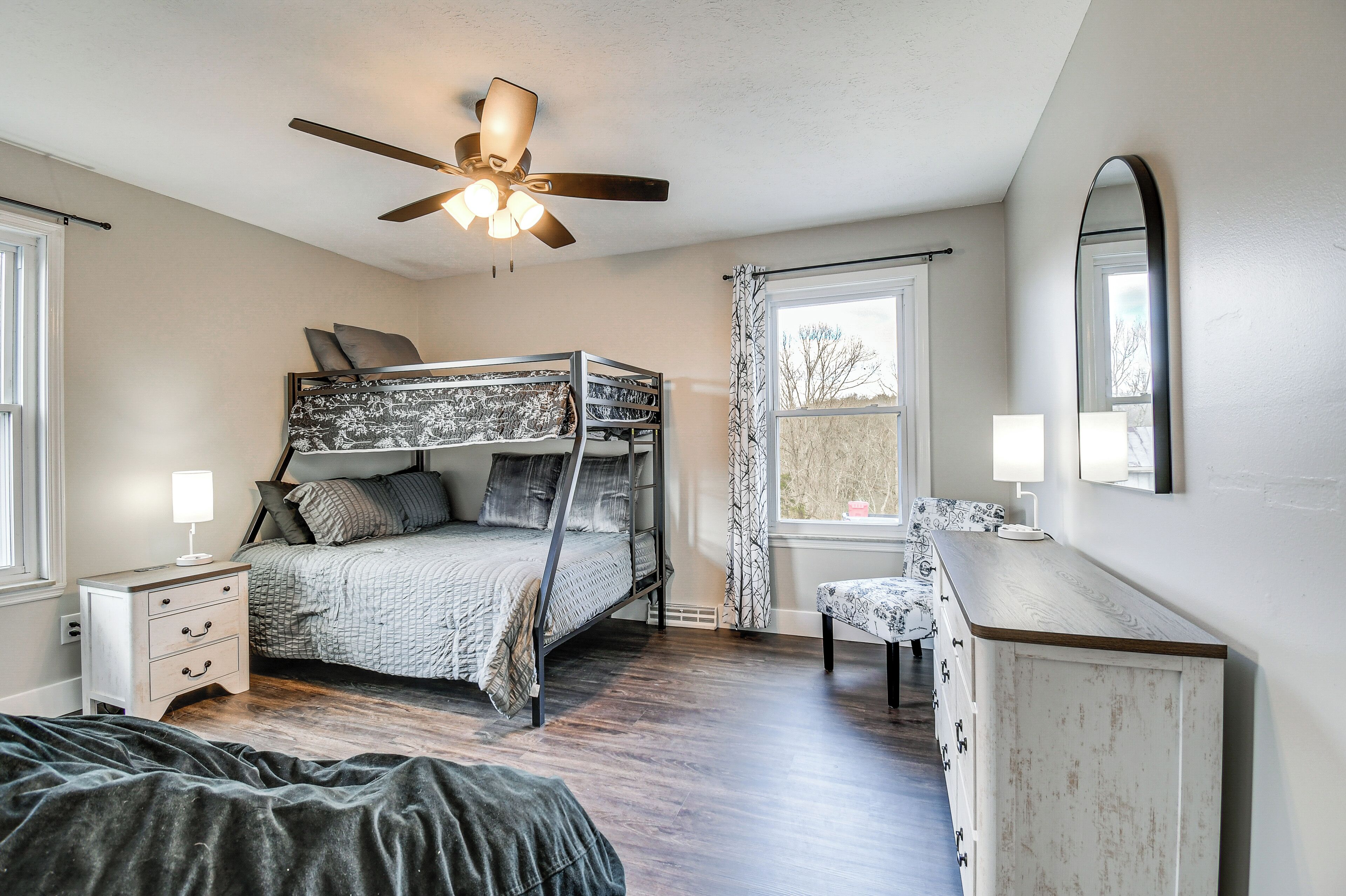 A bedroom with a bunk bed, a bedside table with a lamp, a dresser, a ceiling fan, and a window with curtains.