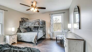 A bedroom with a bunk bed, a bedside table with a lamp, a dresser, a ceiling fan, and a window with curtains.