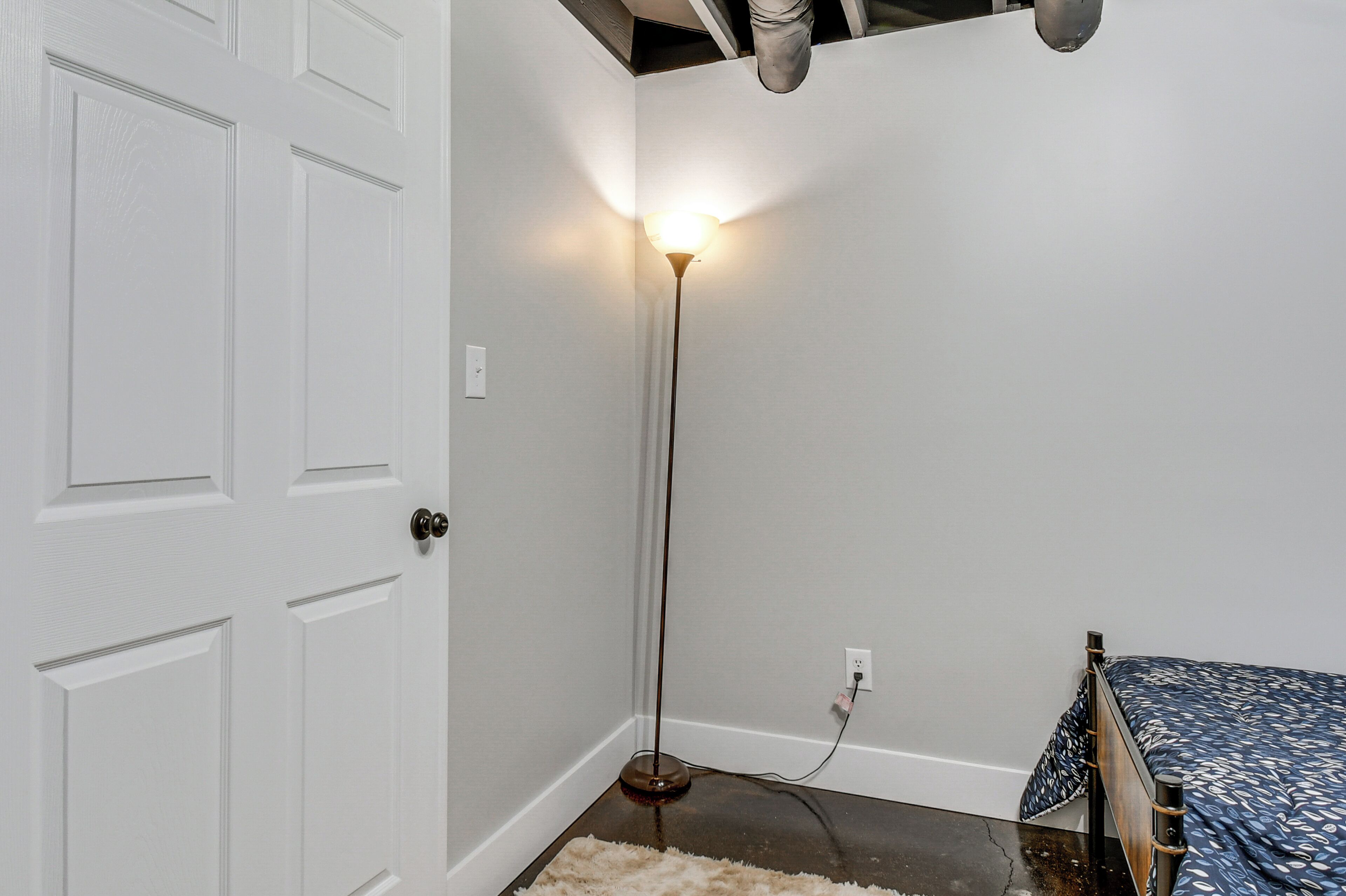 A room with a white door, a standing floor lamp, a bed with a patterned bedspread, and a white wall.