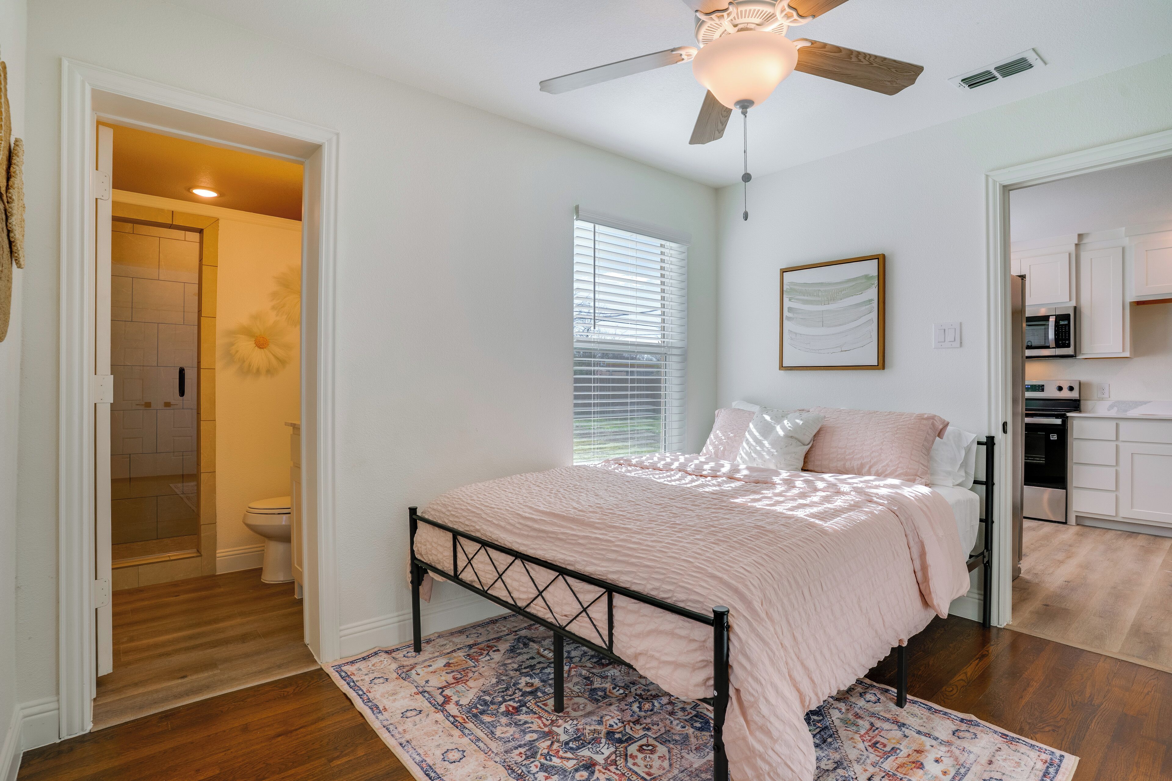 A bedroom with a bed, a ceiling fan, a window with blinds, and a view of the kitchen.