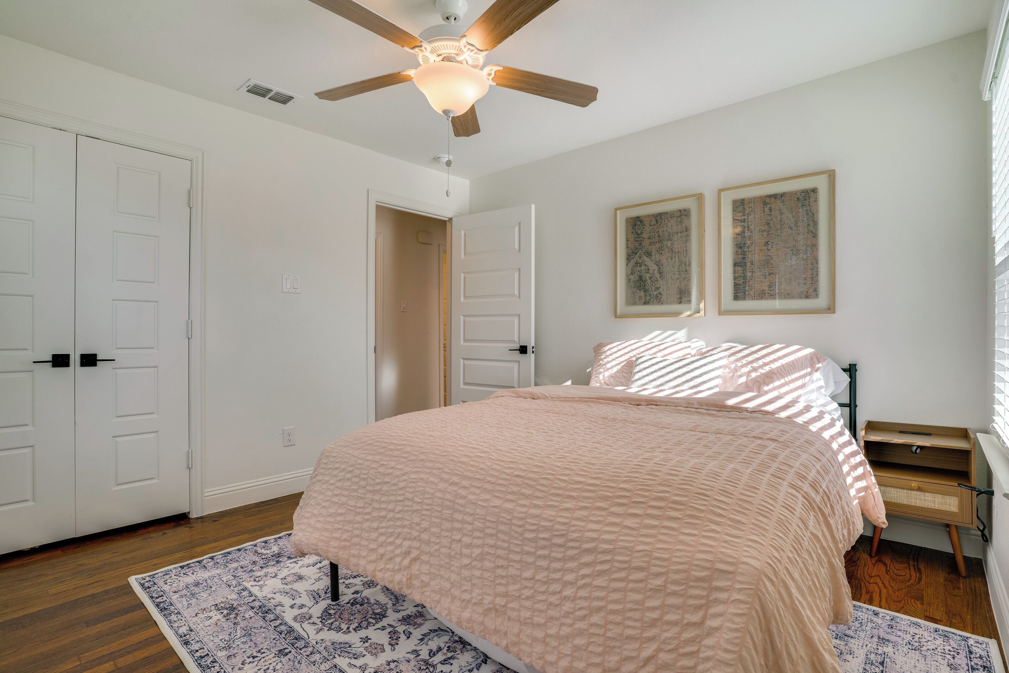 A bedroom with a bed, a ceiling fan, two framed artworks, a nightstand, and a door.