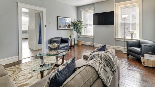 Un salon comprenant un canapé, un fauteuil et une table basse. On y trouve également un ventilateur de plafond, un téléviseur fixé au mur et une fenêtre avec des stores.