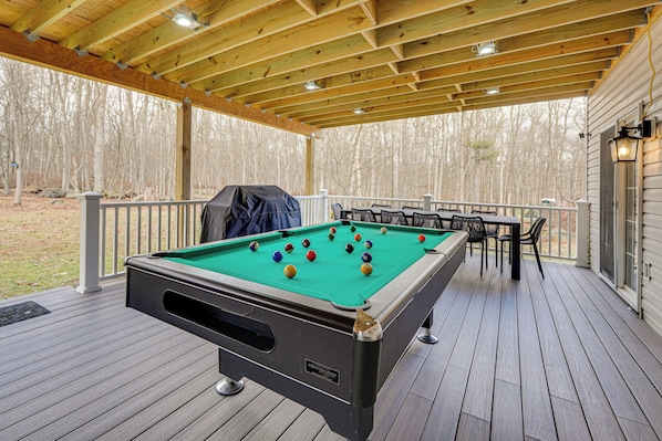 Une table de billard avec des billes posées sur une terrasse en bois, entourée de chaises et d’une table, le tout sous un toit en bois.