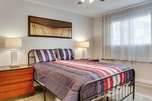 A neatly arranged bedroom with a metal bed frame, a wooden nightstand, two bedside lamps, and a large abstract painting on the wall. - < 1 Mi to Dtwn Hilo: Comfy Condo w/ Pool Access (Hilo)