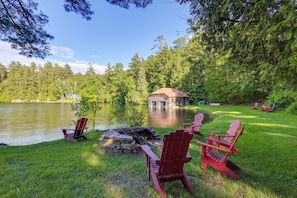 A lakeside cabin with a fire pit, red Adirondack chairs, and a view of the lake and forest. - Expansive Eagle Lake Escape w/ Boathouse + Beach (Ticonderoga)