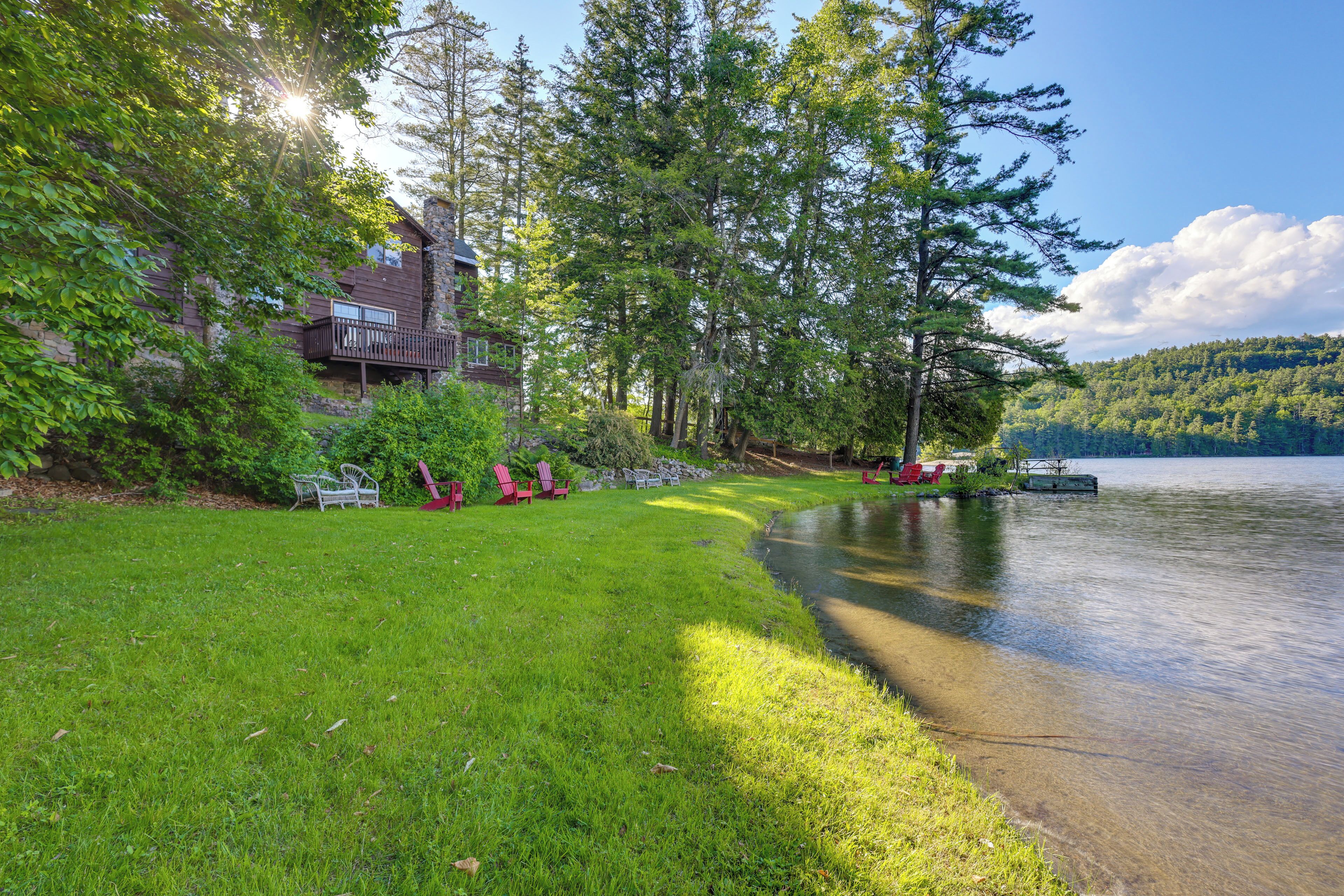 A lakeside cabin with a deck, surrounded by lush greenery and a clear view of the lake.