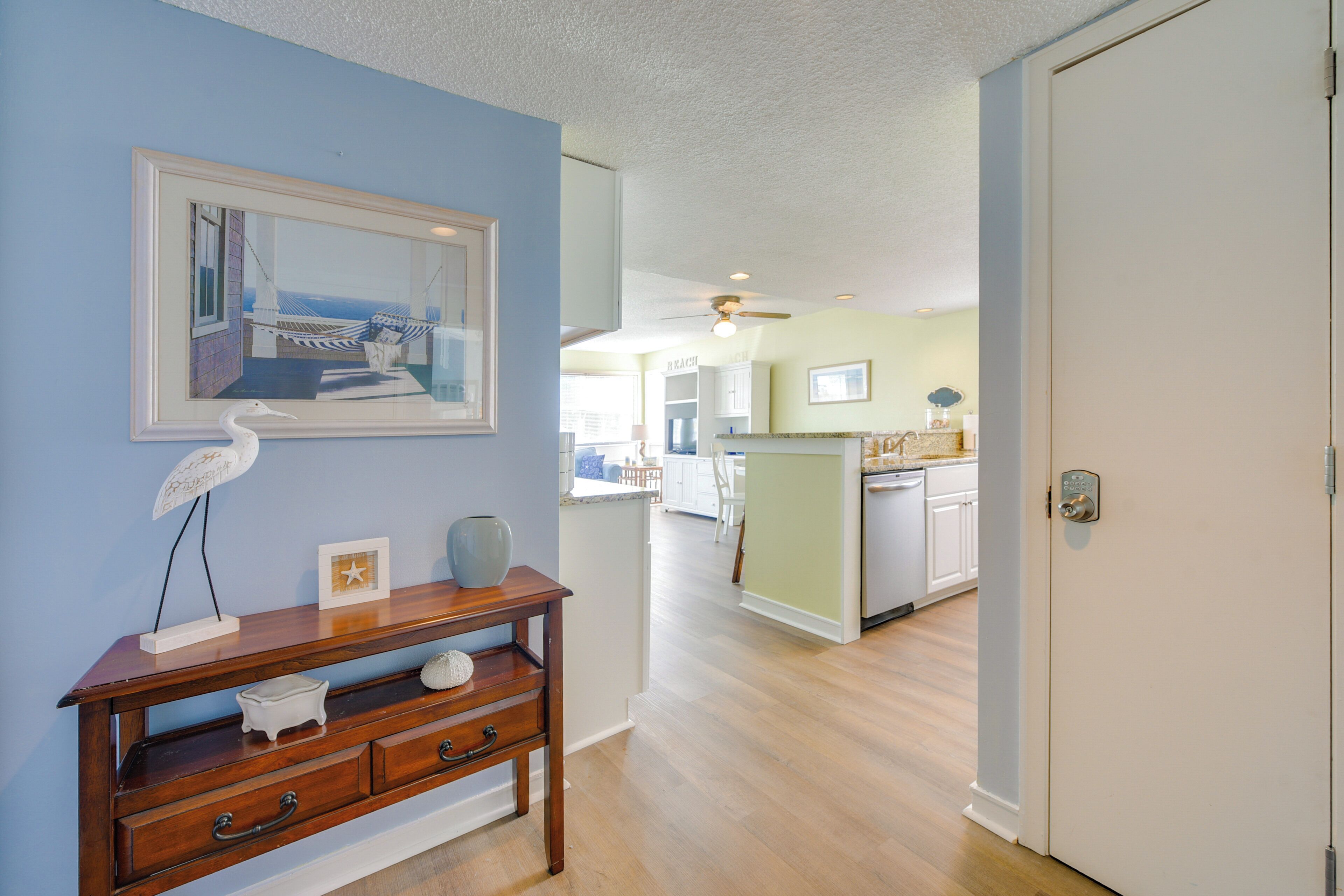 A hallway with a wooden console table, a framed picture, and a white door leading to a kitchen area.