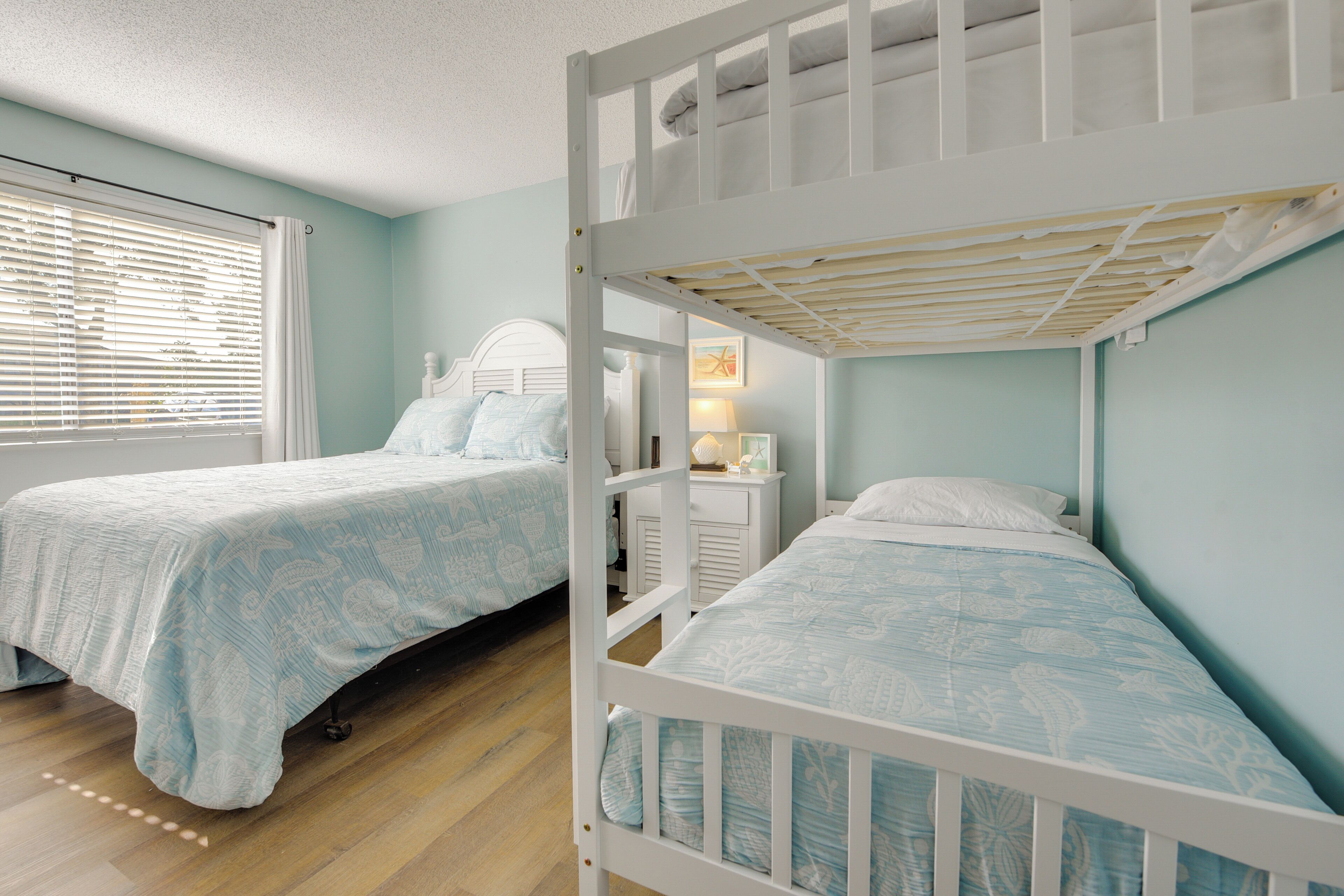 A room with two bunk beds, a window with blinds, a bedside table with a lamp, and a wall-mounted shelf.