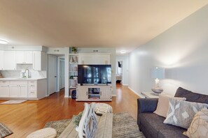 Una sala de estar moderna con un sofá gris, un televisor de pantalla plana y una cocina con armarios blancos y una mesa de comedor.