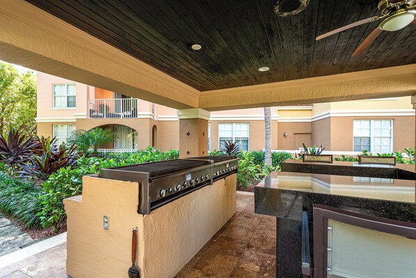 Un espace cuisine extérieur doté d’un barbecue, entouré de plantes et avec un bâtiment en arrière-plan.