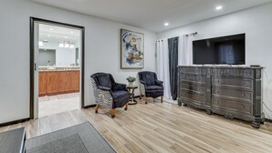 Un salon moderne doté d’un téléviseur à écran plat, de deux fauteuils, d’une commode en bois foncé et d’une cuisine visible à travers une porte ouverte.