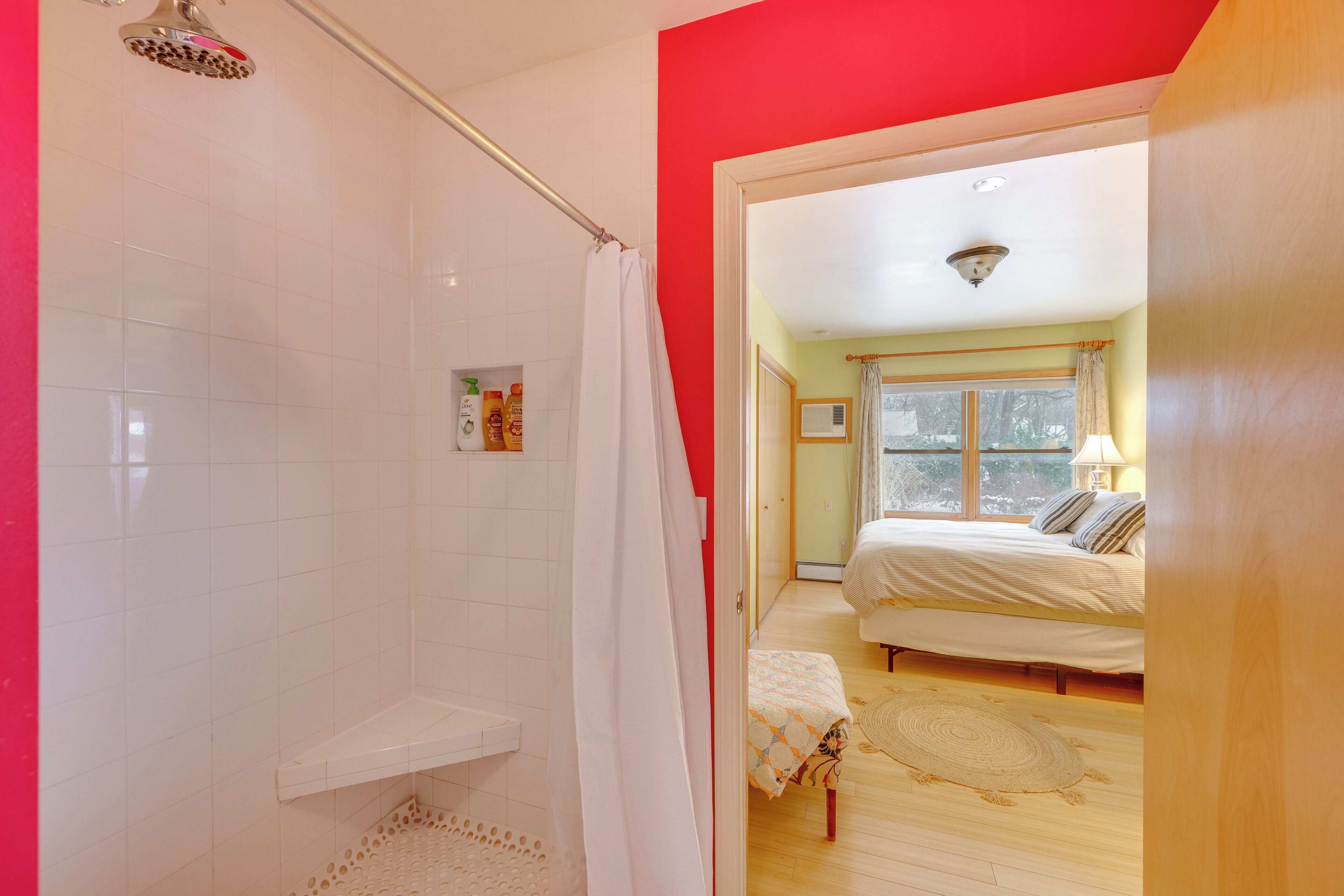 A bathroom with a shower, a toilet, and a sink. There is a bed with a window and a lamp in the adjacent room.