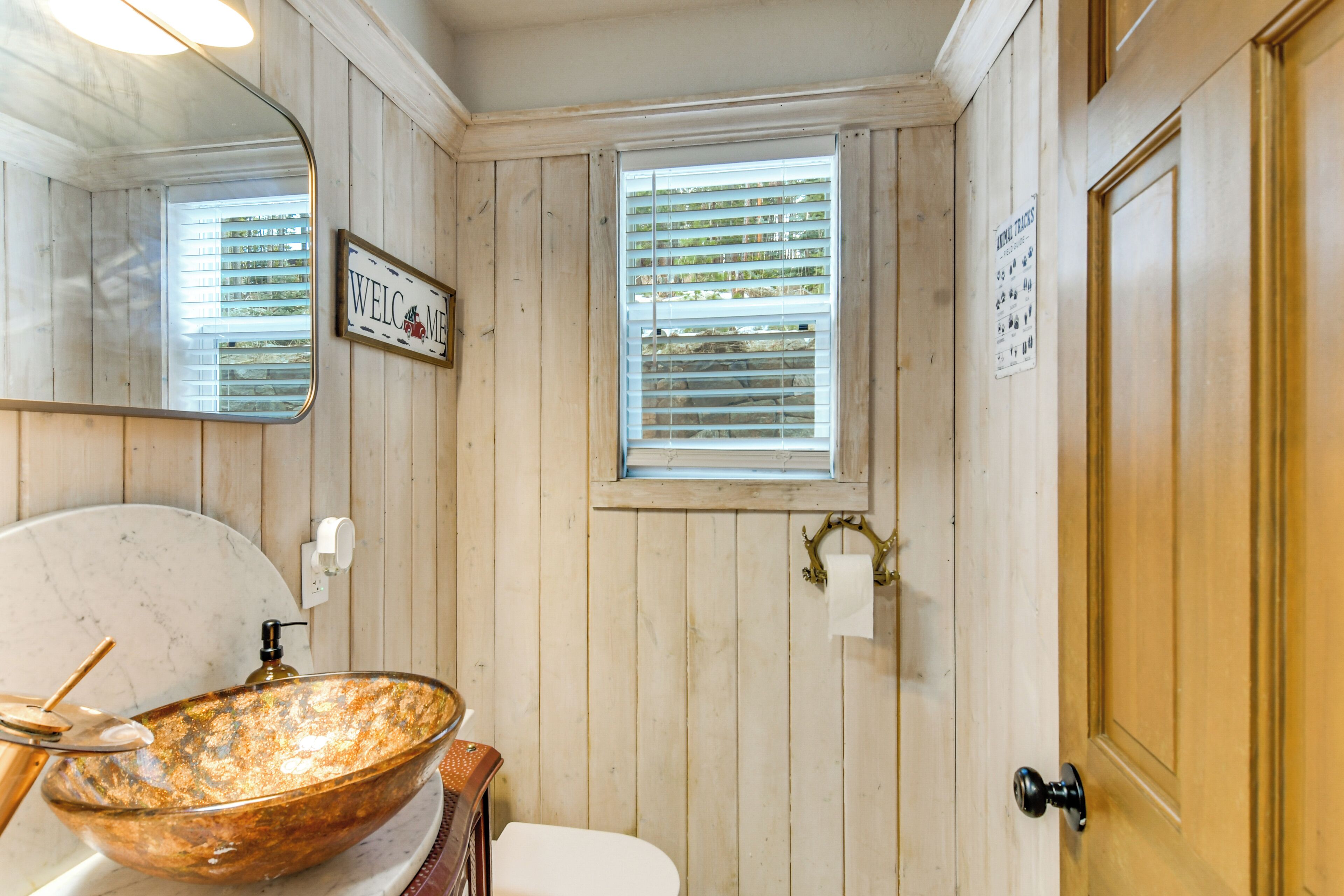 Une salle de bain avec des murs en bois, un lavabo blanc avec un récipient en cuivre, un miroir, une fenêtre avec des stores et une porte avec une pancarte.