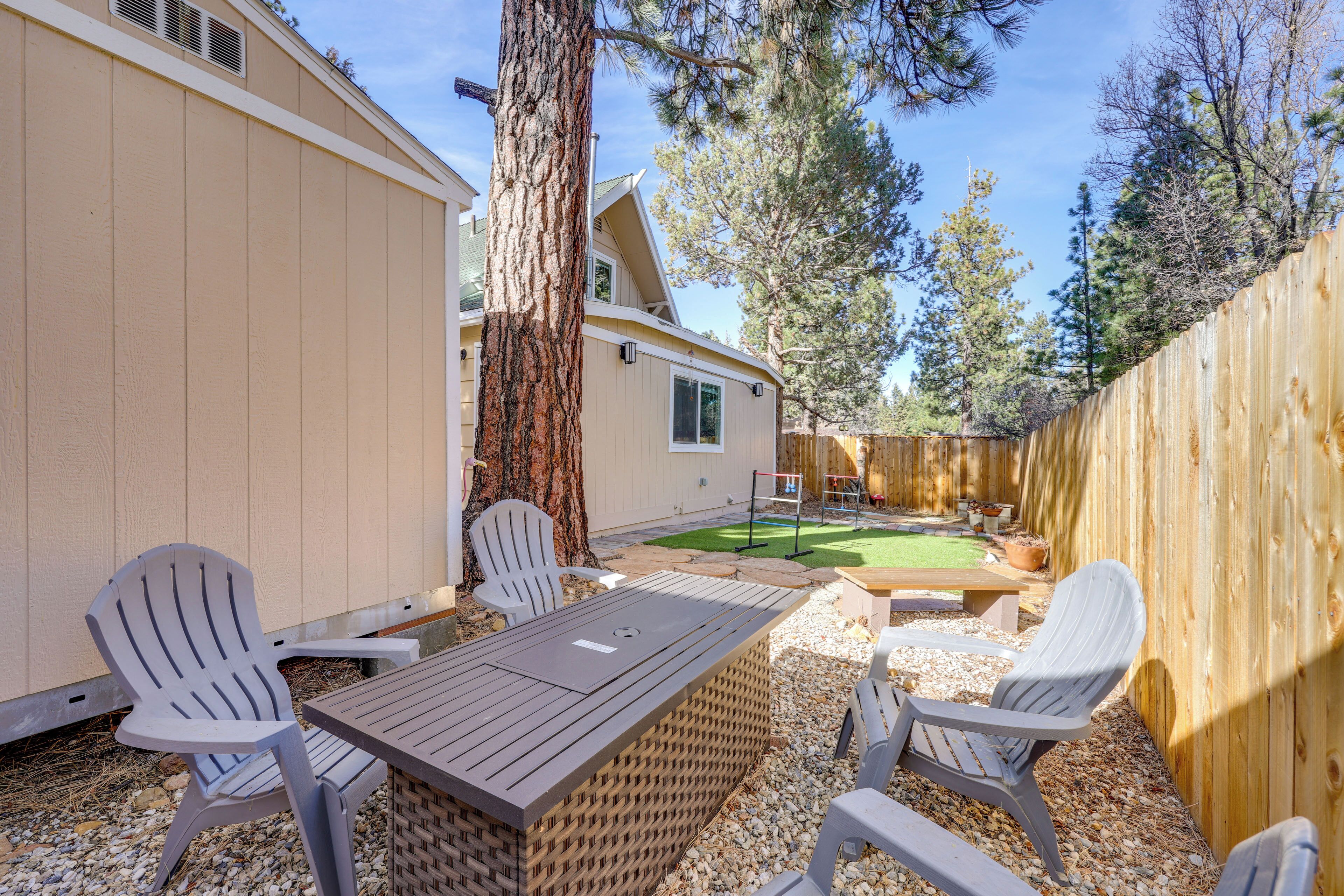 A patio area with a wooden table and four Adirondack chairs, surrounded by a wooden fence and a large tree.