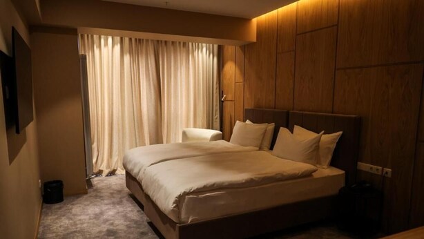 Une chambre d’hôtel avec un grand lit, une tête de lit en bois, une table de chevet et une fenêtre avec des rideaux.