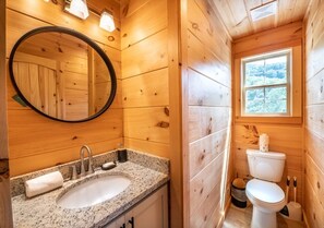A wooden cabin bathroom with a granite sink, a round mirror, a toilet, and a window with a view of trees. - Heated indoor pool | Hot tub | Game Room (Sevierville)