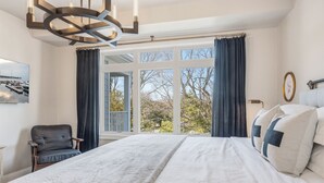 Een slaapkamer met een groot bed, een stoel, een raam met gordijnen en uitzicht op bomen en een steiger.