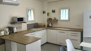 Una cuina moderna amb armaris blancs, un taulell de fusta, microones, bullidor d'aigua i cafetera. Hi ha dues finestres amb cortines, una cadira blanca i una taula petita.
