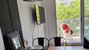 Een eetgedeelte met een houten tafel, zwarte stoelen, een zwarte hanglamp, een ingelijst schilderij en een balkon met een rode stoel.