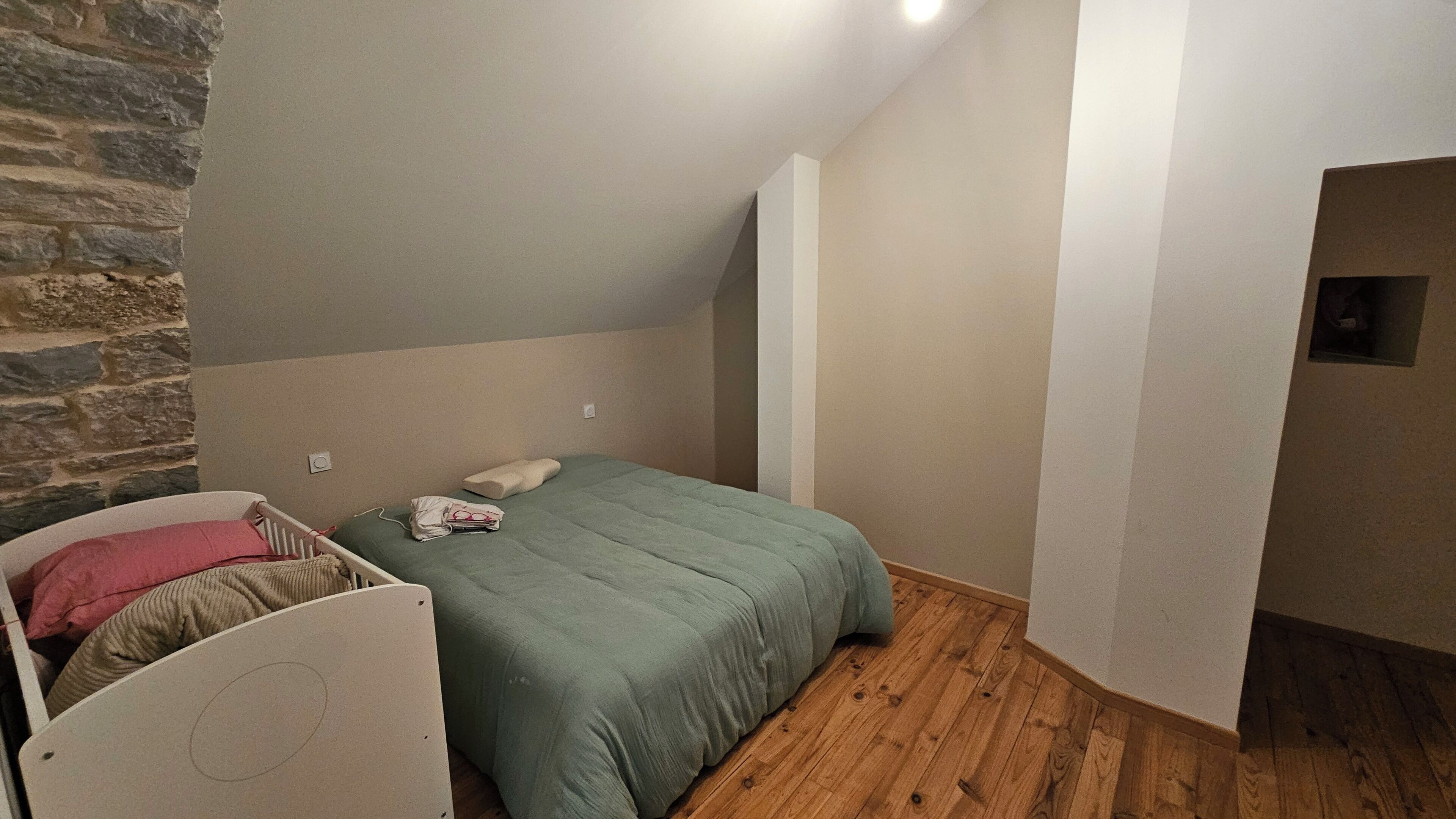 Camera da letto con soffitto spiovente, un letto con copripiumino verde, una testiera bianca e un muro in pietra.