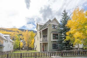 A two-story house with a balcony, surrounded by a wooden fence, set in a residential area with autumn-colored trees. - Box Canyon Basecamp 4 Bedroom Home (Telluride)