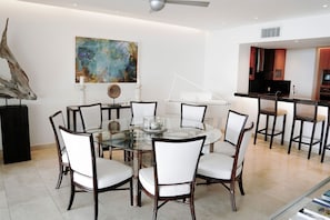 A modern dining area with a round glass table, white upholstered chairs, and a contemporary art piece on the wall. - Portico 1 3 Bedroom Condo (Prospect)