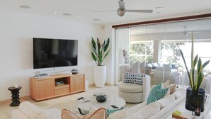 Una sala de estar moderna con un televisor de pantalla plana, un mueble de madera para entretenimiento, una planta grande en maceta, un conjunto de sofás y una mesa de centro de vidrio.