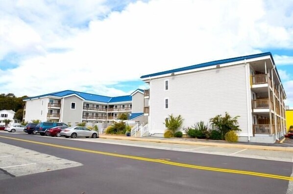 Un motel de varios pisos con techo azul y revestimiento blanco, que cuenta con balcones y un estacionamiento con varios autos.