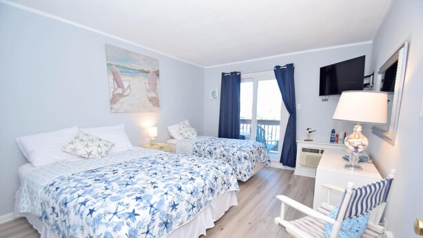 A bedroom with two beds, a desk, a chair, a TV, and a balcony with curtains. - Orleans Court 304 1 Bedroom Condo (Ocean City)