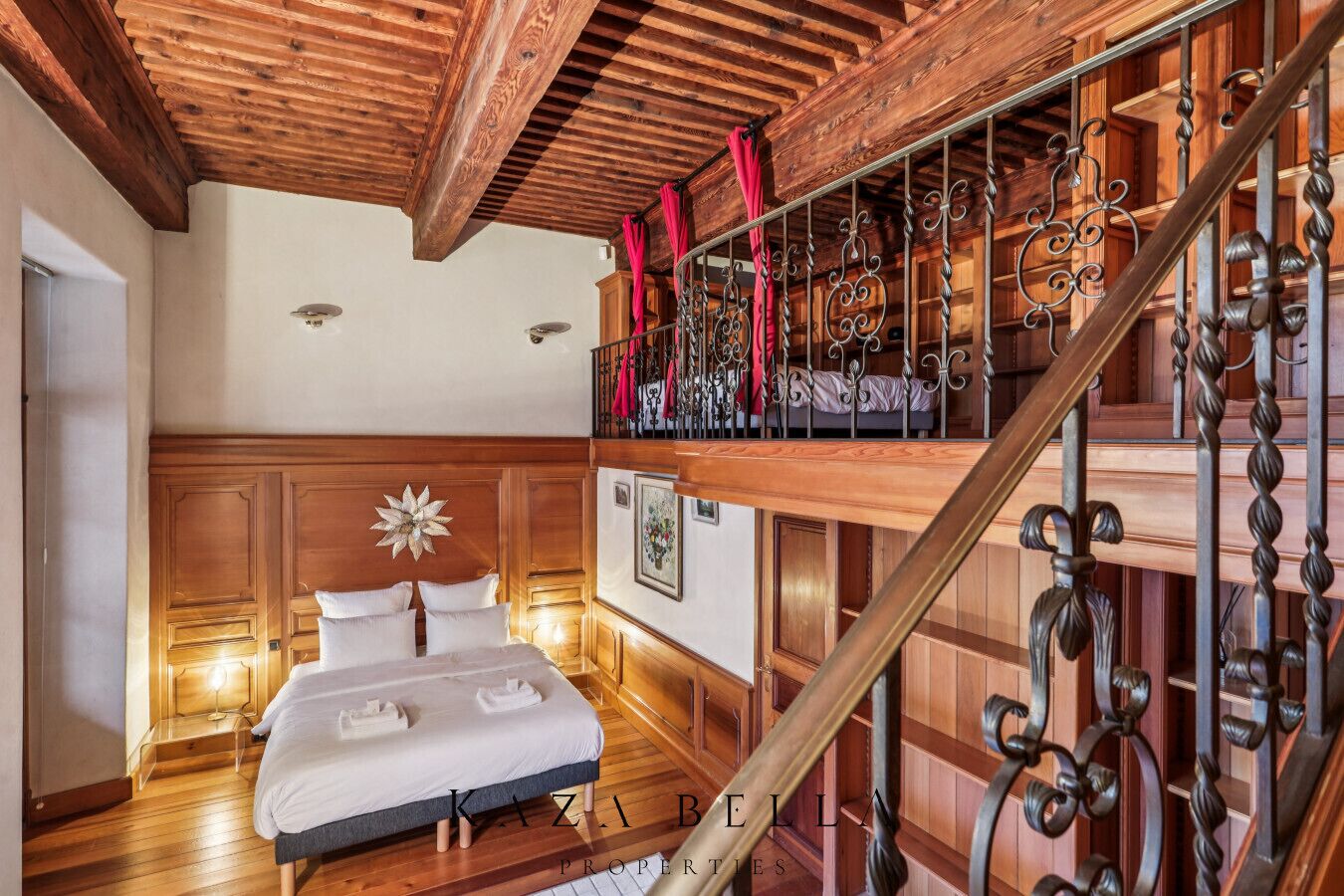 A bedroom with a wooden ceiling, a bed with white linens, a decorative wall piece, and a staircase with wrought iron railings.
