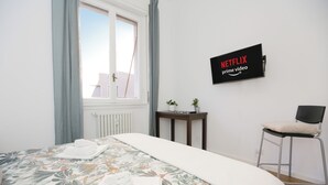 A neatly made bed with floral bedding, a wall-mounted television displaying 'NETFLIX prime video', a small table with two potted plants, and a chair with a cushion. - Centro-Brera, MM2 Moscova-Garibaldi, Corso Como. (Milano)