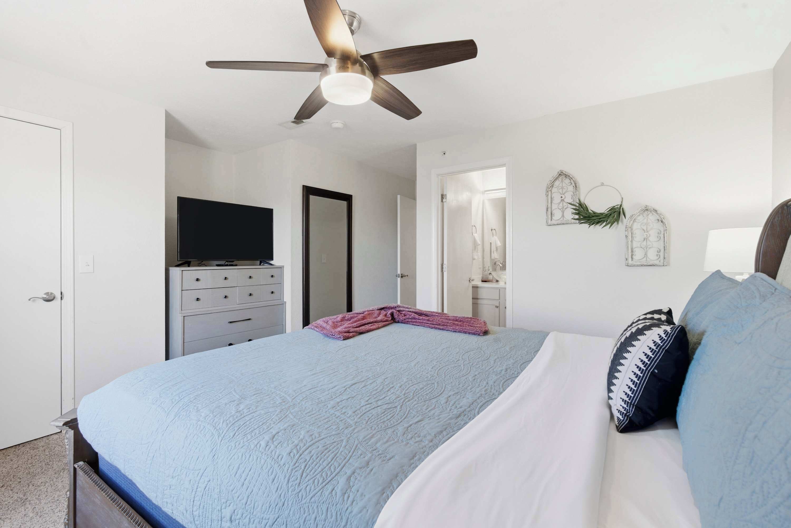 A bedroom with a bed, a ceiling fan, a dresser, a television, and a door leading to another room.