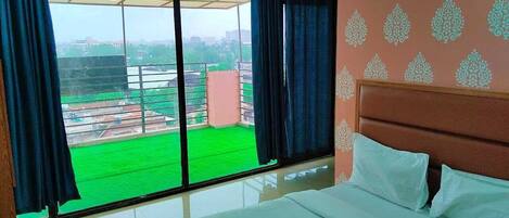 A hotel room with a large window, a bed, and a view of a green field and buildings.