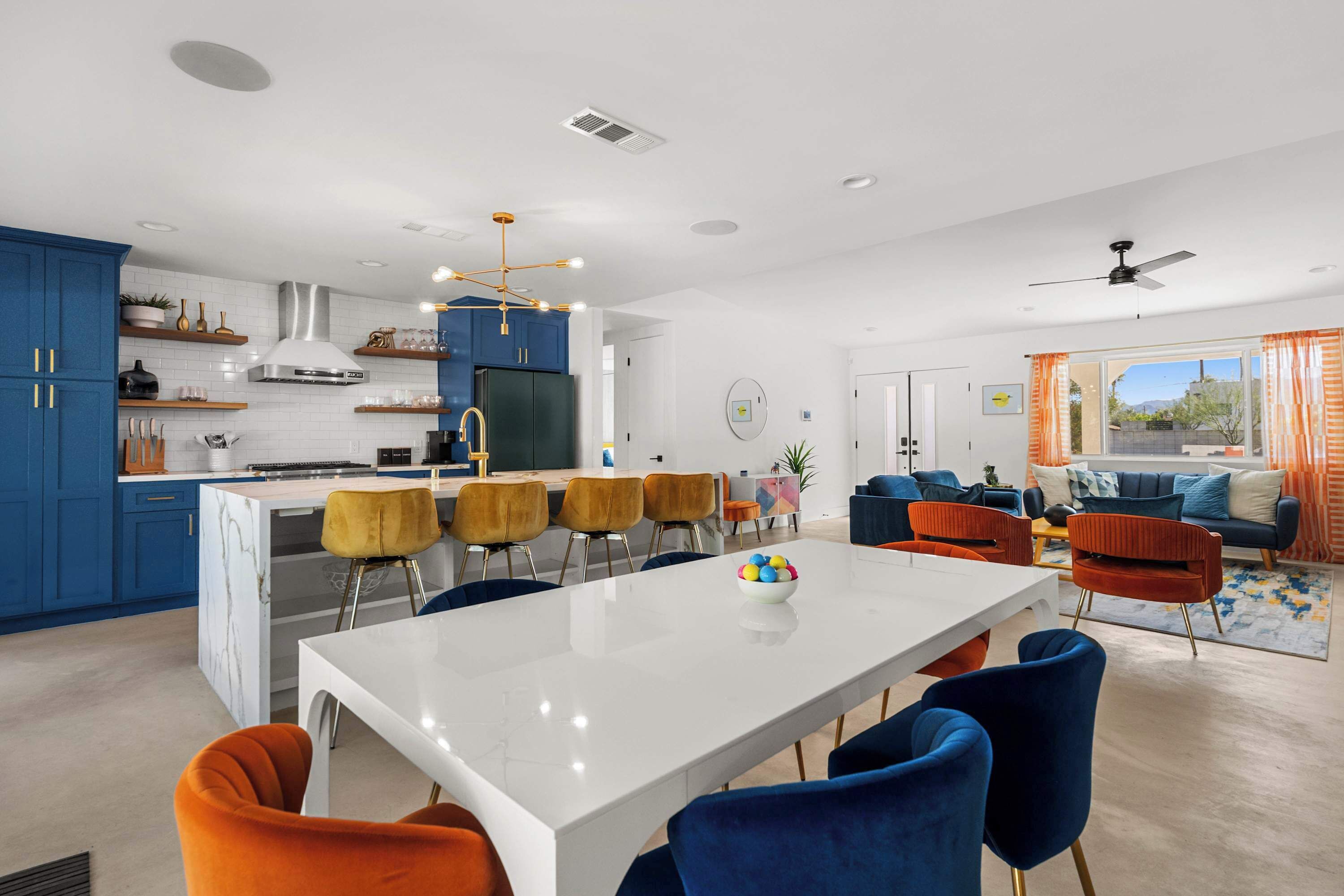 A modern kitchen with blue cabinets, a white island, and a dining area with orange and blue chairs.