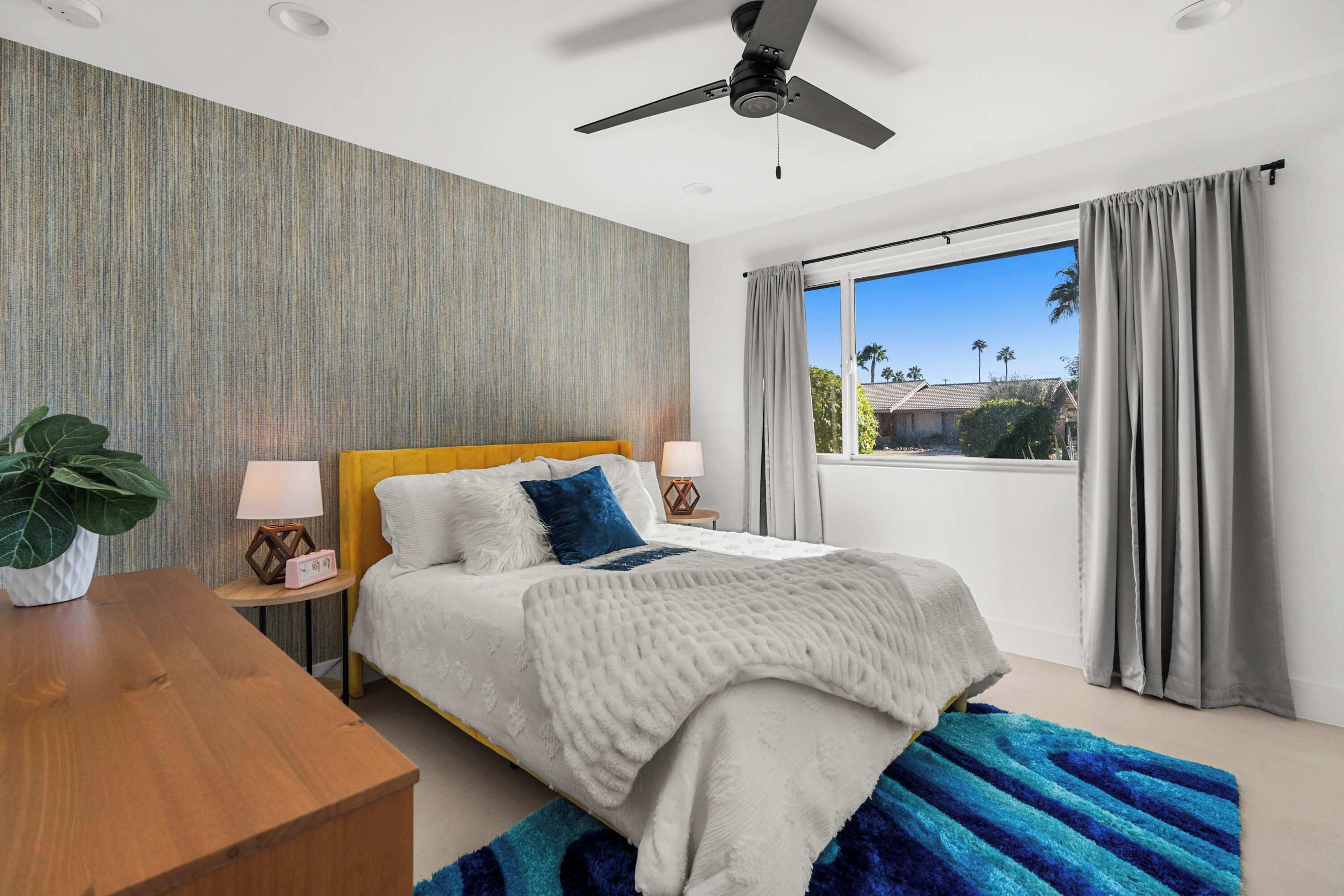 A modern bedroom with a bed, bedside tables, a ceiling fan, a large window with curtains, and a view of palm trees.