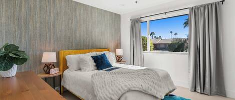A modern bedroom with a bed, bedside tables, a ceiling fan, a large window with curtains, and a view of palm trees.