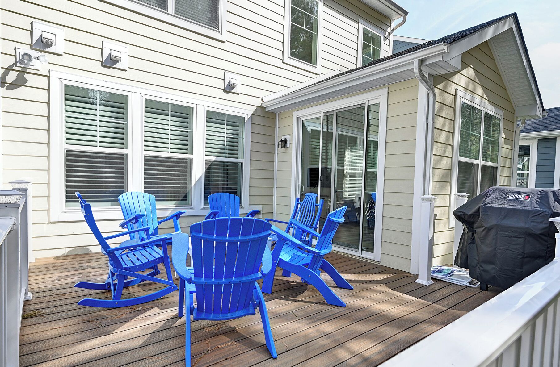 Una terraza con sillas Adirondack azules, una parrilla y una casa de revestimiento blanco con múltiples ventanas.