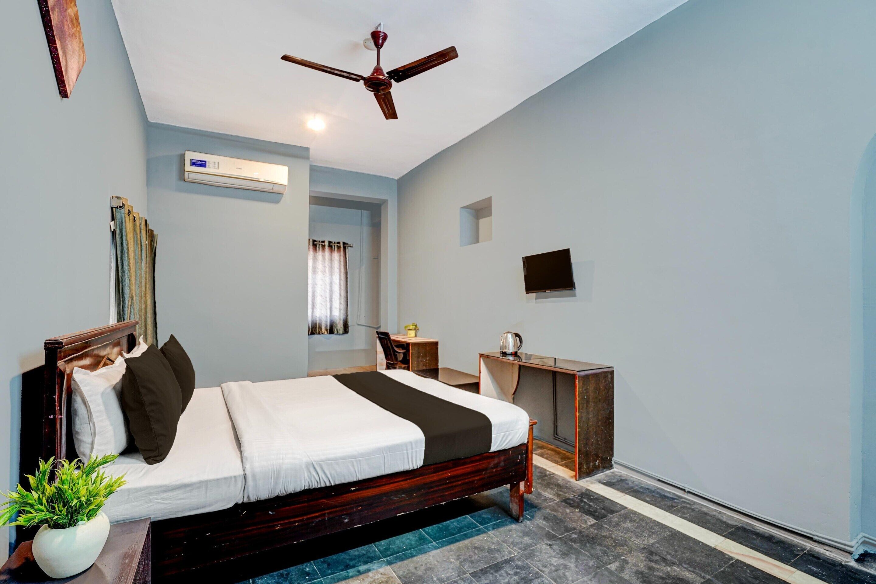 Un dormitorio con una cama, ventilador de techo, aire acondicionado, televisor, escritorio y una planta.