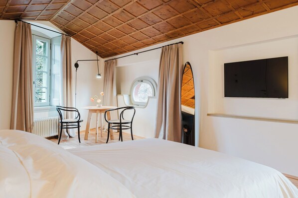 Une chambre avec un grand lit, une petite table ronde avec des chaises, une fenêtre avec des rideaux, un plafond avec des poutres en bois et un téléviseur fixé au mur.