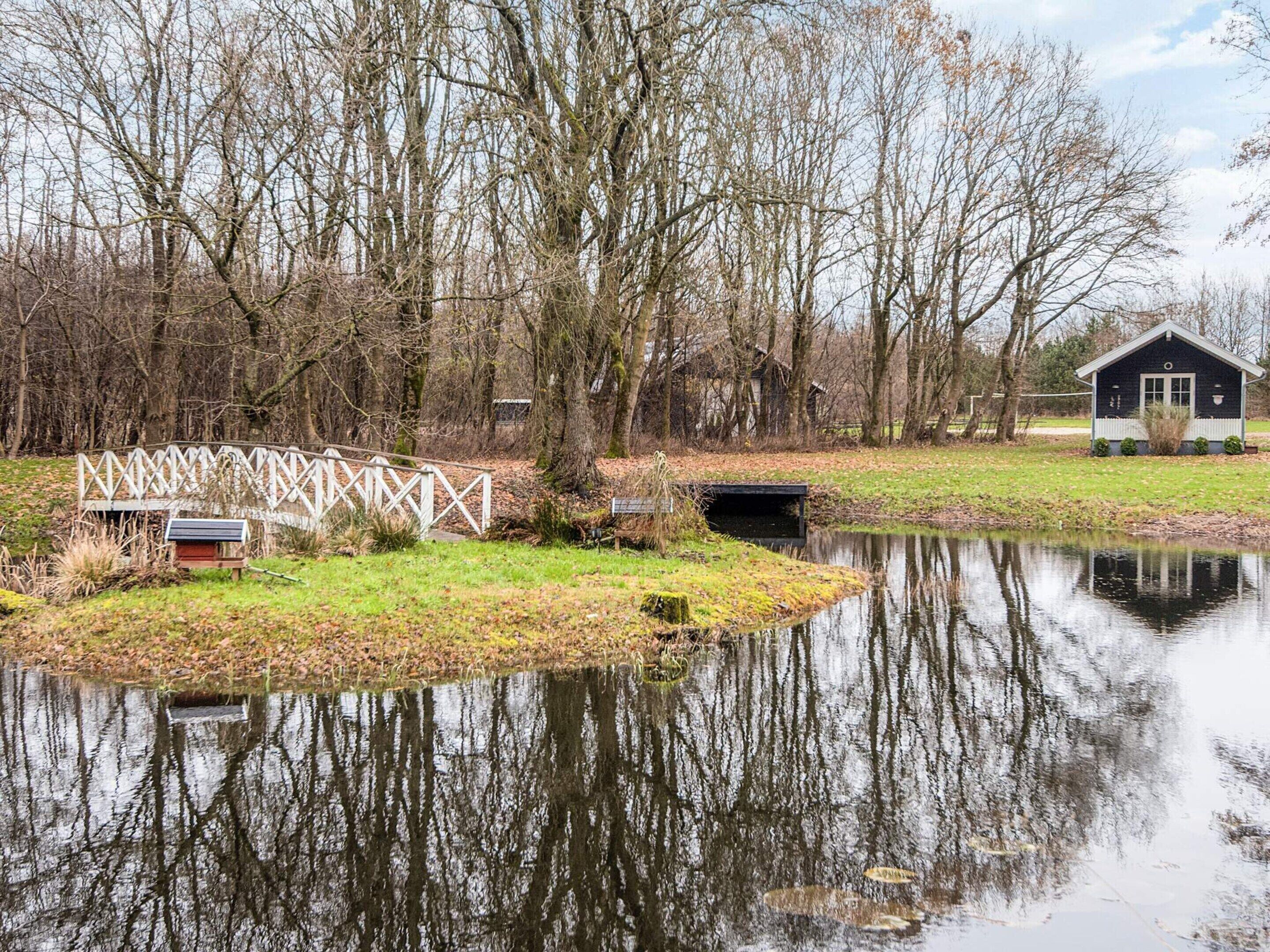 A tranquil lakeside scene with a wooden bridge, a small house, and leaf-covered ground.
