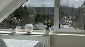 Ett sovrum med ett sluttande tak, en säng med ett grått täcke, ett fönster med utsikt över träd och hus, och två krukväxter på fönsterbrädan.