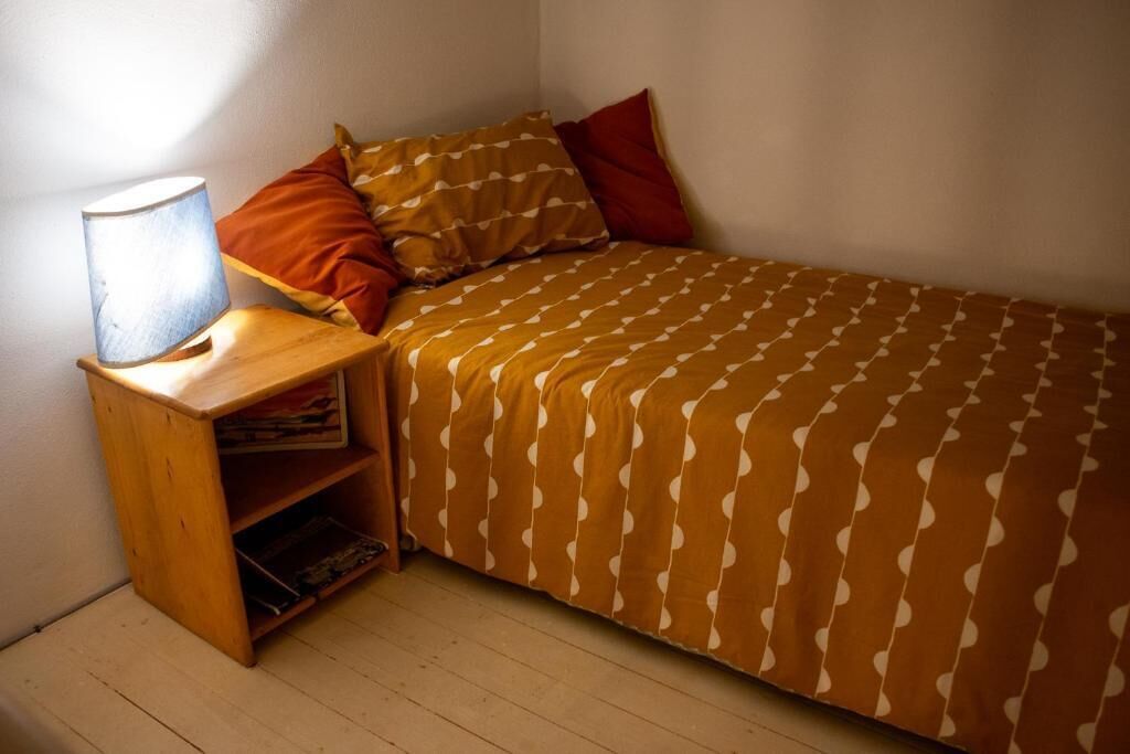 A bedroom with a bed, a nightstand with a lamp, and a shelf with books.