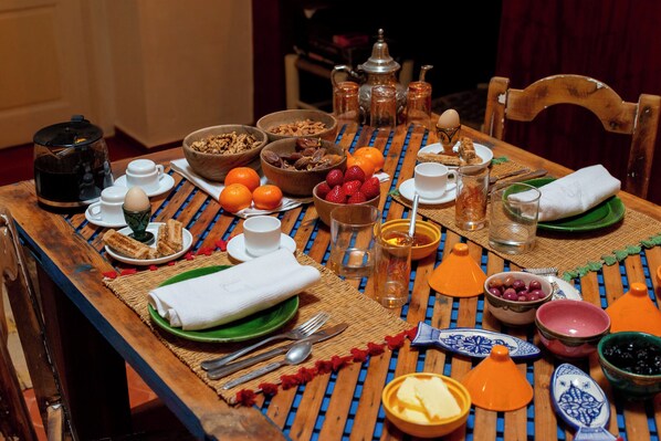 Une table à manger dressée avec différents plats, notamment des fruits, du pain et une théière, ce qui laisse supposer qu’un repas va être servi.