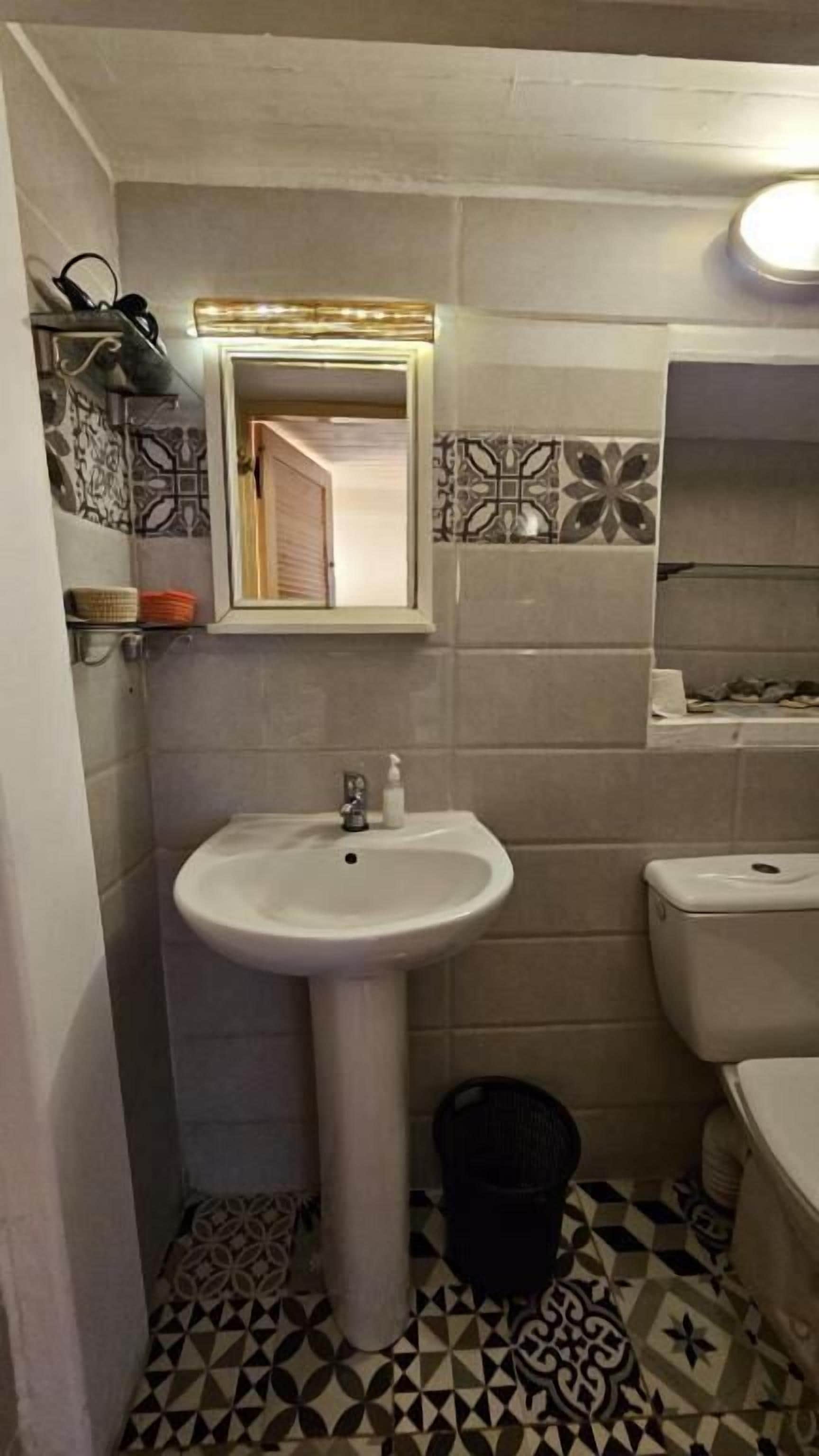 Un baño con un lavamanos blanco, un espejo y un inodoro. El piso presenta un diseño de baldosas geométricas.